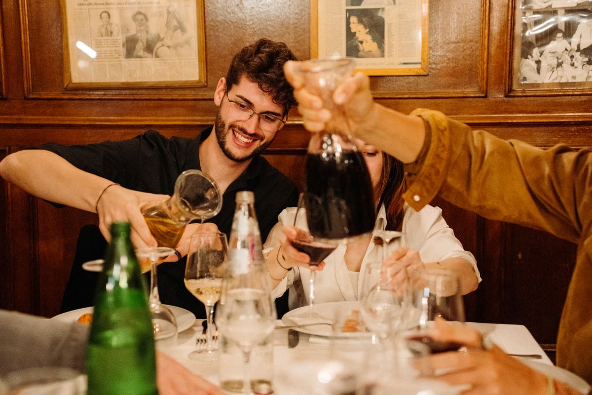 A person holding a craft of wine as two people in the background pour red and white wine.