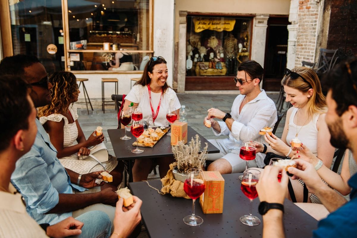 various people sitting at tables enjoying a red beverage and speaking in the Italian language. 