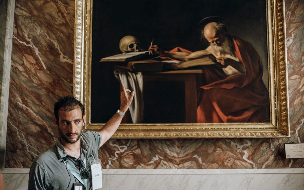 man pointing out art work of old man with skull on table